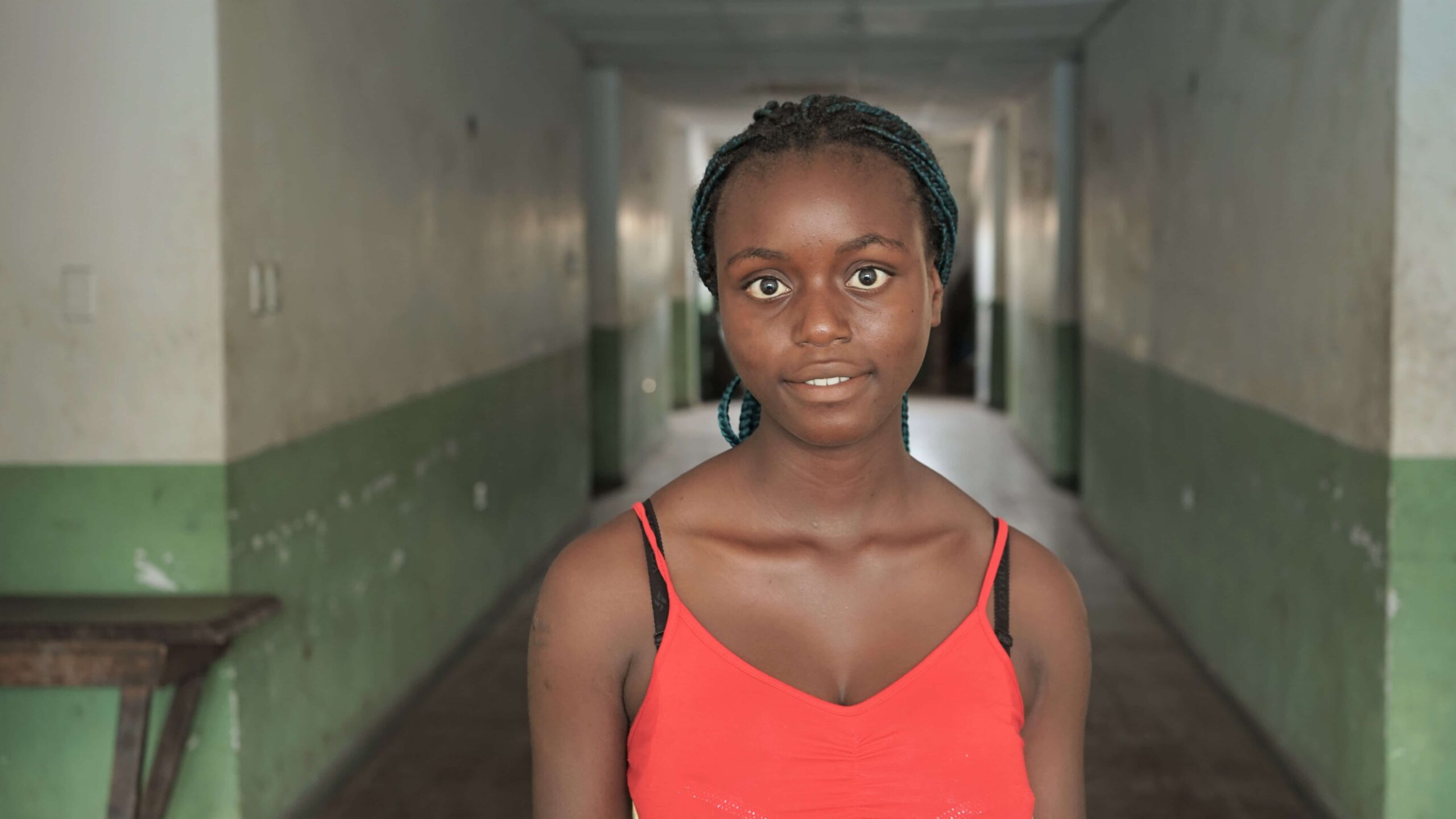Lydia stands in a long corridor wearing an orange blouse and looks into the camera