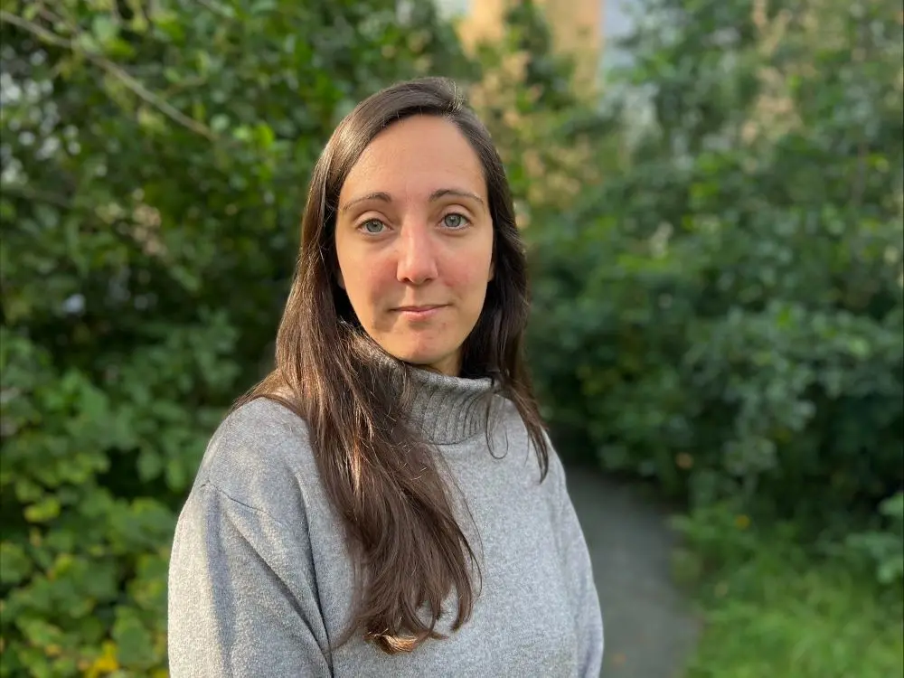 Natasha Bracq, wearing a grey sweater in front of a green bush