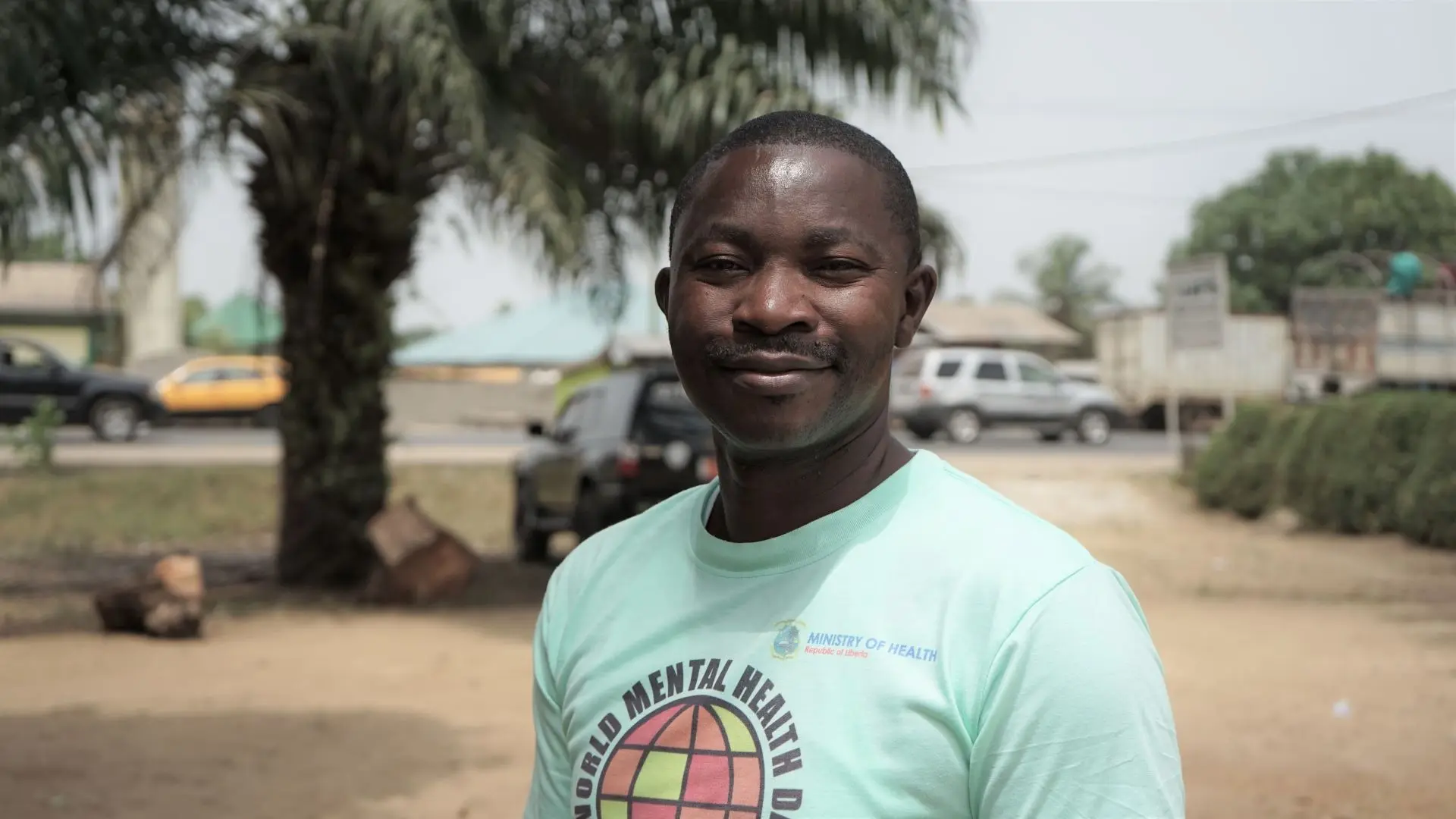 Swaray in blue t-shirt smiling in front of a palm tree in Liberia